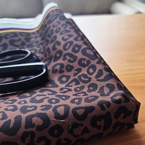 Peut inclure: Un morceau de tissu plié avec un imprimé léopard marron et des taches noires. Une paire de ciseaux noirs repose sur le tissu. Le tissu est sur une surface en bois.