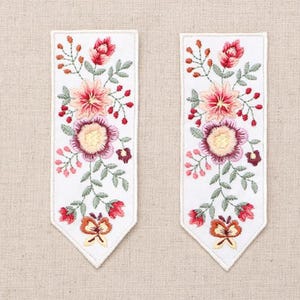 May include: Two white embroidered bookmarks with floral designs. Each bookmark features a central arrangement of pink, red, and yellow flowers, green leaves, and a butterfly at the bottom. The embroidery is detailed and colorful.