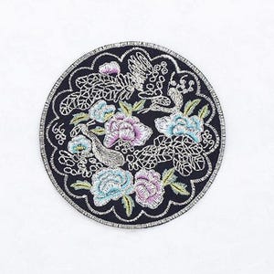 May include: Circular embroidered patch featuring a floral design with birds. The patch has a black background with colorful flowers in shades of blue, purple, and pink. The edges are outlined with a silver thread.