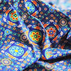 Puede incluir: Primer plano de una tela azul vibrante con un patrón floral repetido. El diseño presenta motivos circulares con acentos amarillos, naranjas y verdes, rodeados de flores más pequeñas y detalles en espiral. La tela está doblada, mostrando su textura.