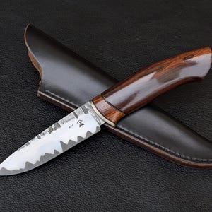 May include: A handcrafted knife with a dark wooden handle and a serrated blade, resting on a black leather sheath. The blade has a unique, textured finish. The knife is designed for outdoor use.