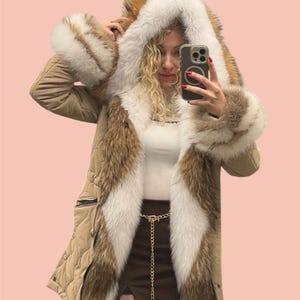 May include: A tan winter coat with a fur-lined hood and cuffs. The coat has a quilted design, a zip closure, and brown fur trim. The fur is a mix of brown, white, and tan colours. A gold chain belt is visible.