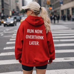 May include: A red hoodie with the text "RUN NOW. OVERTHINK LATER." in white. The hoodie is worn by a person with blonde hair, a beige cap, and sunglasses, standing on a crosswalk in a city.