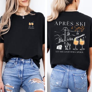 May include: Black t-shirt with a small graphic of two cocktails and the name "Christin" on the front. The back features a mountain scene with the words "Apr&egrave;s Ski & Spritz" and "Let the good times aperol."