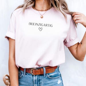 Könnte beinhalten: Ein hellrosa T-Shirt mit kurzen Ärmeln, das den schwarzen Schriftzug (W)EINZIGARTIG und ein kleines Herzsymbol darunter aufweist. Das T-Shirt wird mit einer blauen Jeans und einem braunen Ledergürtel mit Metallschnalle sowie einer Goldkette getragen.