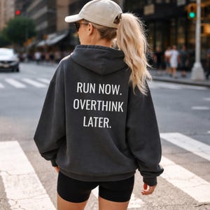 May include: A charcoal gray hoodie with the text "RUN NOW. OVERTHINK LATER." in white block letters. The hoodie is worn with black shorts and a beige baseball cap. The person is standing on a crosswalk in a city.