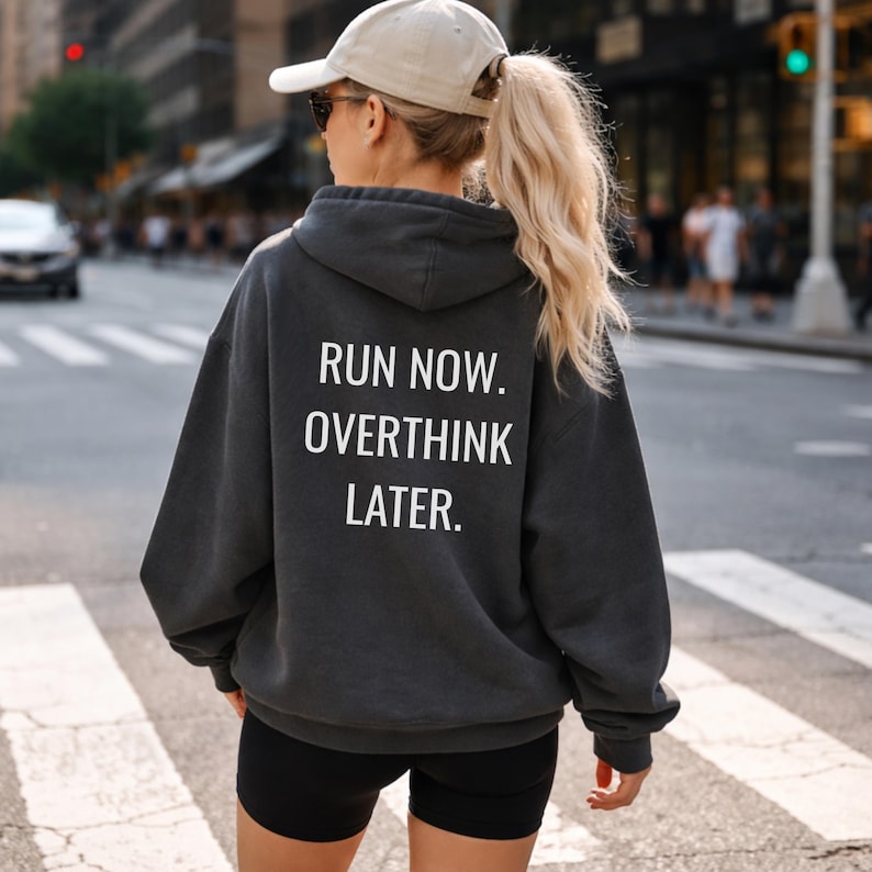 May include: A charcoal gray hoodie with the text "RUN NOW. OVERTHINK LATER." in white block letters. The hoodie is worn with black shorts and a beige baseball cap. The person is standing on a crosswalk in a city.
