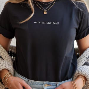 Könnte beinhalten: Schwarzes T-Shirt mit dem weißen Text "MY KIDS HAVE PAWS". Das Shirt hat hochgekrempelte Ärmel und wird mit einer blauen Jeans kombiniert. Die Person trägt goldene Halsketten und Armbänder.