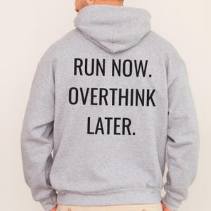 May include: A light gray hoodie with the text "RUN NOW. OVERTHINK LATER." printed in black block letters across the back. The hoodie has a hood and long sleeves. The person wearing the hoodie is facing away from the camera.
