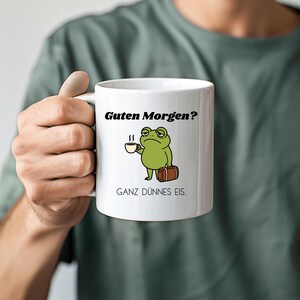 Könnte beinhalten: Ein weißer Keramikbecher mit einer Illustration eines grünen Frosches, der eine kleine Kaffeetasse und einen braunen Koffer hält. Der Text "Guten Morgen?" steht über dem Frosch und der Text "GANZ DÜNNES EIS.." steht unter der Abbildung.