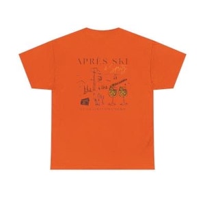 May include: Orange t-shirt with a graphic design on the back. The design includes the words "APRES SKI" above a sketch of a mountain scene, cocktails, and the phrase "IT'S ALWAYS COCKTAIL HOUR".