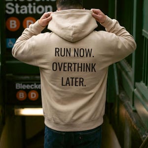 May include: Tan hoodie with the text "RUN NOW. OVERTHINK LATER." printed in black. The hoodie has a hood and long sleeves. The person is wearing blue jeans. The background shows a subway station.