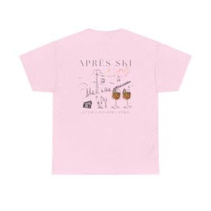May include: A light pink t-shirt featuring a graphic design with the words "Apr&egrave;s Ski" and a winter scene with ski lifts, a cabin, and two glasses of a beverage. The shirt is made of a soft material.