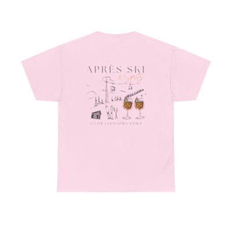 May include: A light pink t-shirt featuring a graphic design with the words "Apr&egrave;s Ski" and a winter scene with ski lifts, a cabin, and two glasses of a beverage. The shirt is made of a soft material.