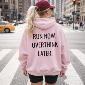 May include: A light pink hoodie with the text "RUN NOW. OVERTHINK LATER." in black. The hoodie is worn by a person wearing a maroon baseball cap and black shorts. The person is standing in a crosswalk in a city.