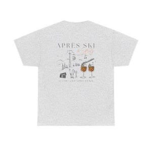 May include: A light gray t-shirt with a graphic design on the back. The design features the words "Apr&egrave;s Ski" above a drawing of a ski resort scene with two cocktails. The text "It's for relaxation time" is below the drawing.
