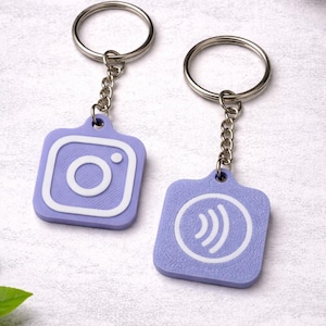 May include: Two keychains with a silver ring and chain. The keychains are lavender-colored, square-shaped, and feature white graphic designs. One has an Instagram logo, and the other has a Wi-Fi symbol.