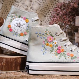 May include: Pair of high-top canvas sneakers in a light green hue, adorned with floral embroidery. One shoe features a Converse All Star logo patch. The embroidered flowers include pink, orange, and yellow blossoms with green leaves.