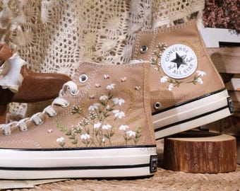 May include: Tan high-top sneakers with white embroidered floral and bee designs. The sneakers feature white laces, a white rubber sole, and the Converse All Star logo patch. The design includes small white flowers and green leaves.