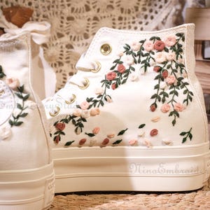 May include: Cream-colored high-top sneakers with floral embroidery. The shoes feature a white platform sole and are adorned with pink and red embroidered flowers and green leaves. The Converse logo is visible on the side.