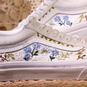 Peut inclure: Baskets en toile blanches avec broderies florales complexes. Le design présente des fleurs bleu clair, des feuilles vertes et des accents marron. Les chaussures ont des lacets et des semelles blancs, avec le texte "NinaEmbroideryStudio".