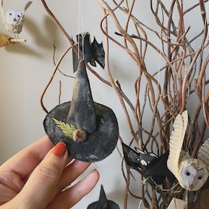 May include: A black witch hat decoration with a small pumpkin and dried leaves attached. The hat is held by a hand, with other Halloween-themed decorations, including a bat, owl, and branches, in the background.
