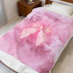 Op de afbeelding: Een roze deken met een grote, lichtroze strik in het midden. De deken heeft een wazige achtergrond met witte spikkels. De deken ligt op een bed met een wit kussen en een houten nachtkastje op de achtergrond.