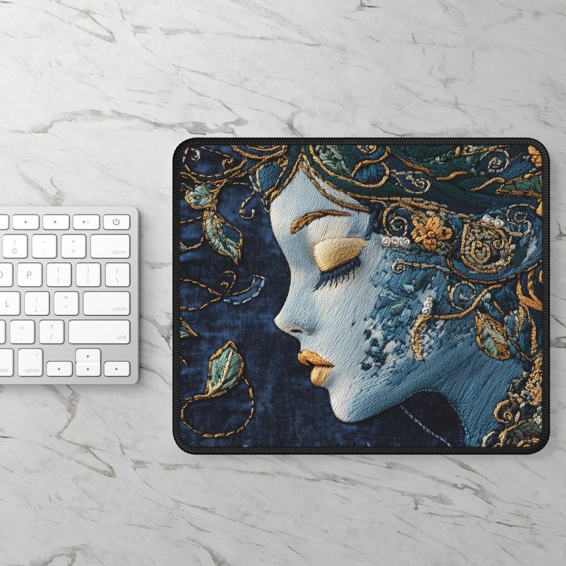 May include: A blue and gold embroidered mouse pad featuring a woman's face with her eyes closed. The woman's face is surrounded by intricate floral designs.