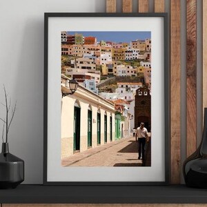 May include: Framed print of a colorful cityscape with buildings of various colors. A man walks down a brick-paved street lined with buildings with green doors and windows. A church tower is visible in the background.