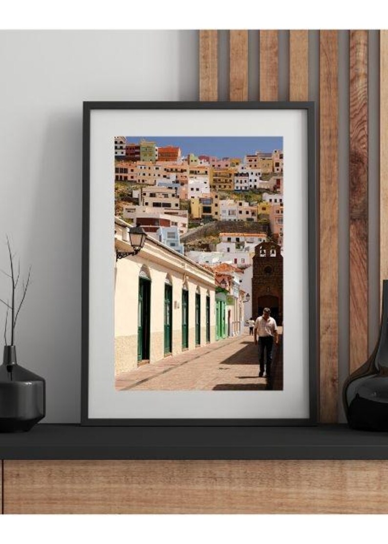 May include: Framed print of a colorful cityscape with buildings of various colors. A man walks down a brick-paved street lined with buildings with green doors and windows. A church tower is visible in the background.