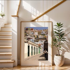 May include: A framed photograph of a street scene in a European town. The image shows a man walking down a cobblestone street lined with buildings. The buildings are painted in various colors, and the sky is blue. The frame is a light wood color.