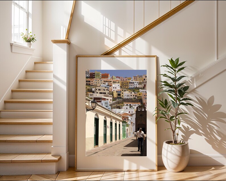 May include: A framed photograph of a street scene in a European town. The image shows a man walking down a cobblestone street lined with buildings. The buildings are painted in various colors, and the sky is blue. The frame is a light wood color.