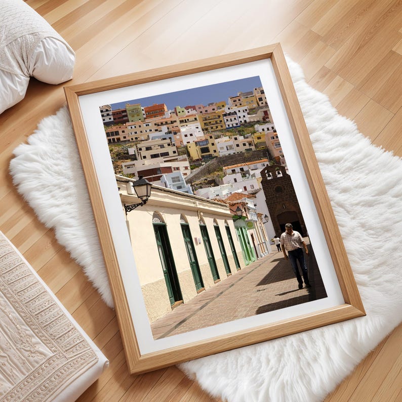 May include: Framed photograph of a colorful hillside town with buildings in various colors. The image is in a light wood frame and is displayed on a white faux fur rug. A person walks down a cobblestone street.