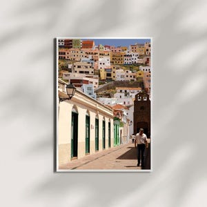 May include: A framed print of a colorful hillside town. The buildings are painted in various colors, including white, yellow, orange, and green. A man walks down a brick-paved street lined with buildings with green doors and windows.