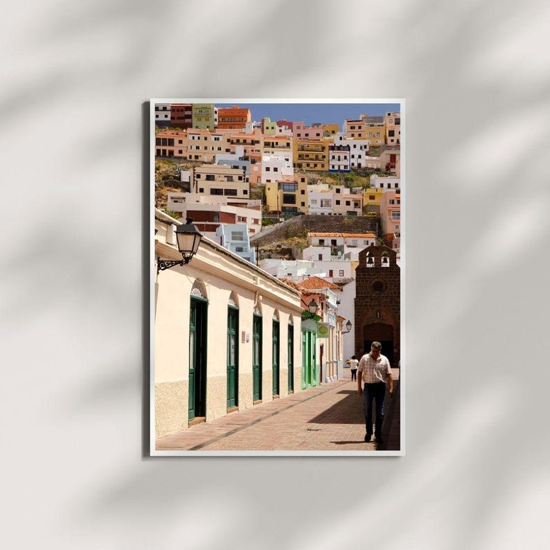 May include: A framed print of a colorful hillside town. The buildings are painted in various colors, including white, yellow, orange, and green. A man walks down a brick-paved street lined with buildings with green doors and windows.