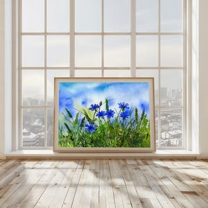 Könnte beinhalten: Aquarellmalerei mit blauen Blumen in einem Feld mit grünem Gras und blauem Himmel. Das Kunstwerk ist in einem hellbraunen Holzrahmen gerahmt. Das Gemälde wird vor einem großen Fenster mit Blick auf die Stadt ausgestellt.