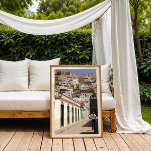 May include: A framed print of a street scene in a European town. The image shows a man walking down a cobblestone street lined with white buildings and colorful houses on a hillside. The print is displayed on a wooden deck.