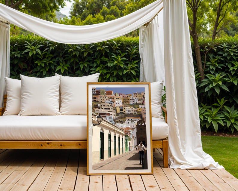 May include: A framed print of a street scene in a European town. The image shows a man walking down a cobblestone street lined with white buildings and colorful houses on a hillside. The print is displayed on a wooden deck.