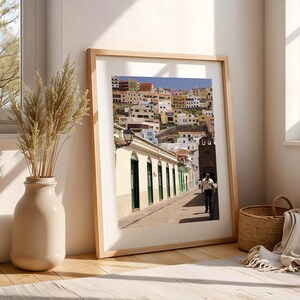 May include: A framed print of a colorful cityscape with buildings of various colors. The print is in a light wood frame and leans against a wall. A vase with dried plants and a woven basket are in the foreground.