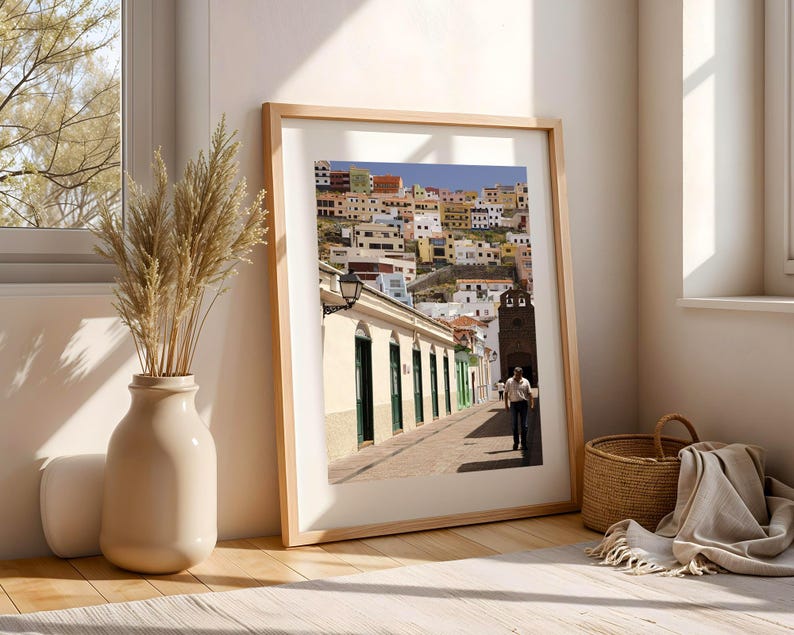May include: A framed print of a colorful cityscape with buildings of various colors. The print is in a light wood frame and leans against a wall. A vase with dried plants and a woven basket are in the foreground.
