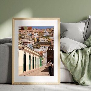 May include: A framed photograph of a street scene in a European town. The image features a man walking down a cobblestone street lined with buildings in various colors. The buildings are white, yellow, and orange with green doors and windows. A church tower is visible.