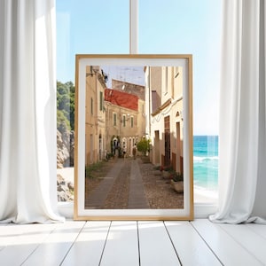 May include: A framed print of a European street scene. The image shows a narrow cobblestone street lined with buildings in shades of beige and cream. A red-tiled roof is visible. The print is displayed in a room with a window overlooking the ocean.