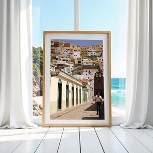 May include: A framed print of a coastal town scene. The image features a narrow street lined with buildings in various colors, leading towards a church tower. A person walks down the street. The frame is a light wood color.