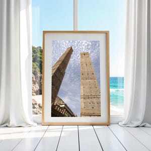 Op de afbeelding: Een ingelijste foto van twee hoge, bruine bakstenen torens tegen een blauwe lucht met witte wolken. De torens leunen lichtjes, waardoor een perspectief ontstaat. De foto is ingelijst in een licht houten lijst.