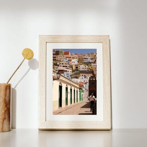 May include: A framed photograph of a street scene in a European town. The image shows a narrow street with buildings on either side, and a person walking down the street. The buildings are painted in various colors, and the sky is blue.