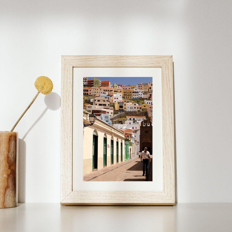 May include: A framed photograph of a street scene in a European town. The image shows a narrow street with buildings on either side, and a person walking down the street. The buildings are painted in various colors, and the sky is blue.