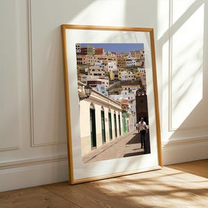 May include: A framed print of a colorful hillside town with buildings in shades of yellow, orange, and white. A street with a person walking is in the foreground. The frame is a light wood color, and the print is leaning against a white wall.