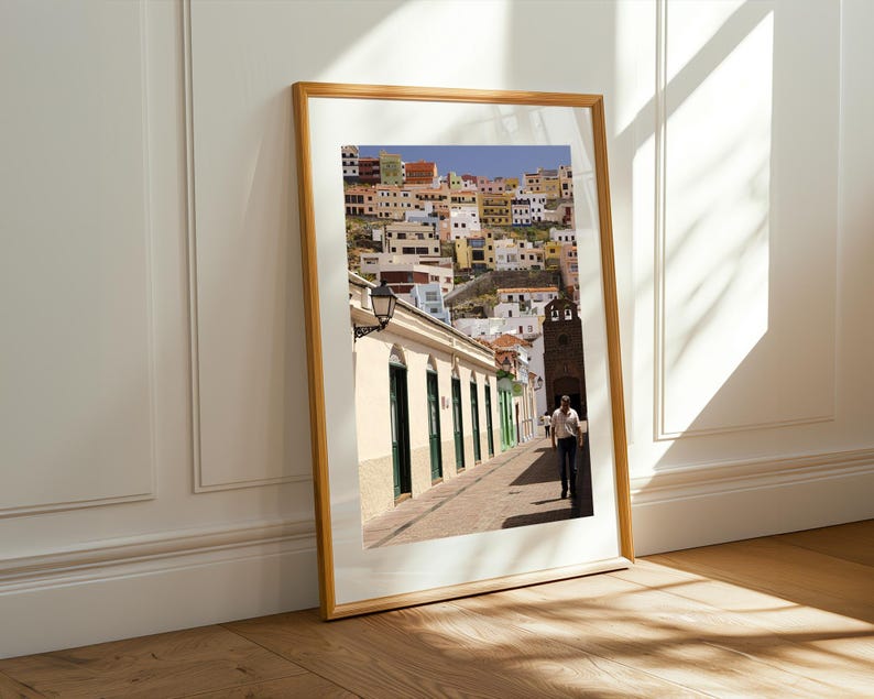 May include: A framed print of a colorful hillside town with buildings in shades of yellow, orange, and white. A street with a person walking is in the foreground. The frame is a light wood color, and the print is leaning against a white wall.