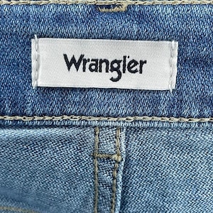 May include: Close-up of blue denim fabric with a white label stitched on, bearing the black word "Wrangler". The denim has visible stitching and a classic jeans design.
