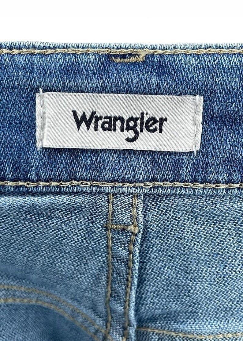 May include: Close-up of blue denim fabric with a white label stitched on, bearing the black word "Wrangler". The denim has visible stitching and a classic jeans design.